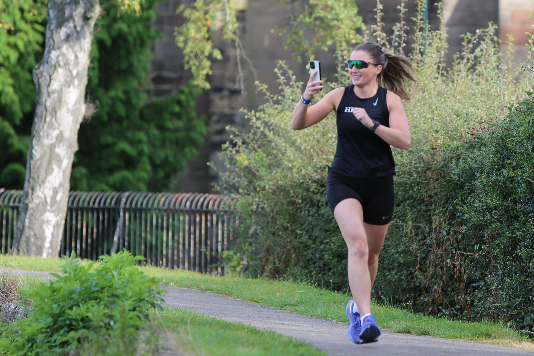 Woman in black athletic wear and sunglasses smiling while running and filming herself with a phone, on a curved park path lined with greenery.