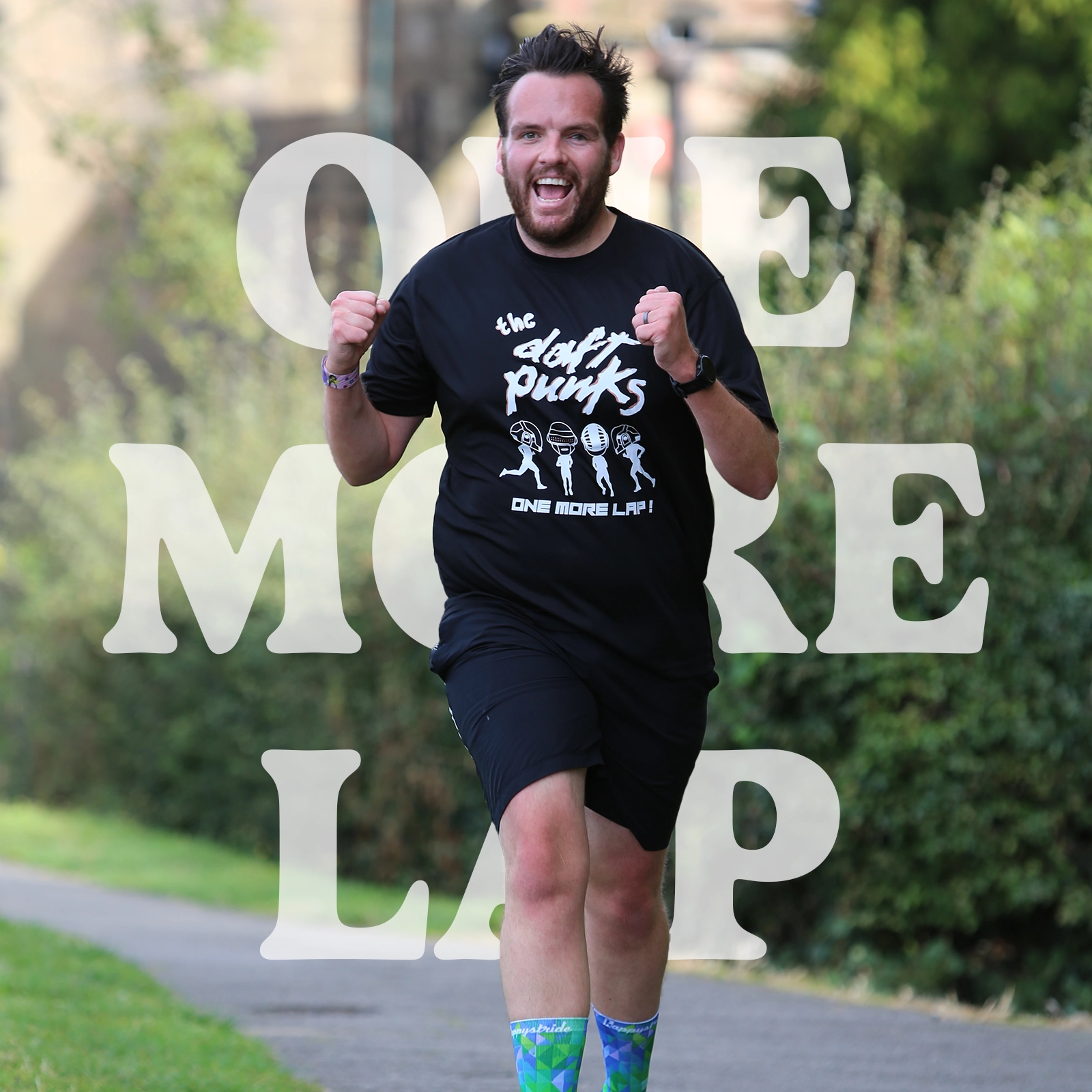 A joyful runner wearing a black t-shirt with the text 'The Dafty Punks - One More Lap!' and a graphic of dancing figures. The runner has a big smile, clenched fists in a celebratory pose, and is running on a paved path surrounded by greenery. The background features a stone wall, and the words 'One More Lap' are overlaid in large white text.
