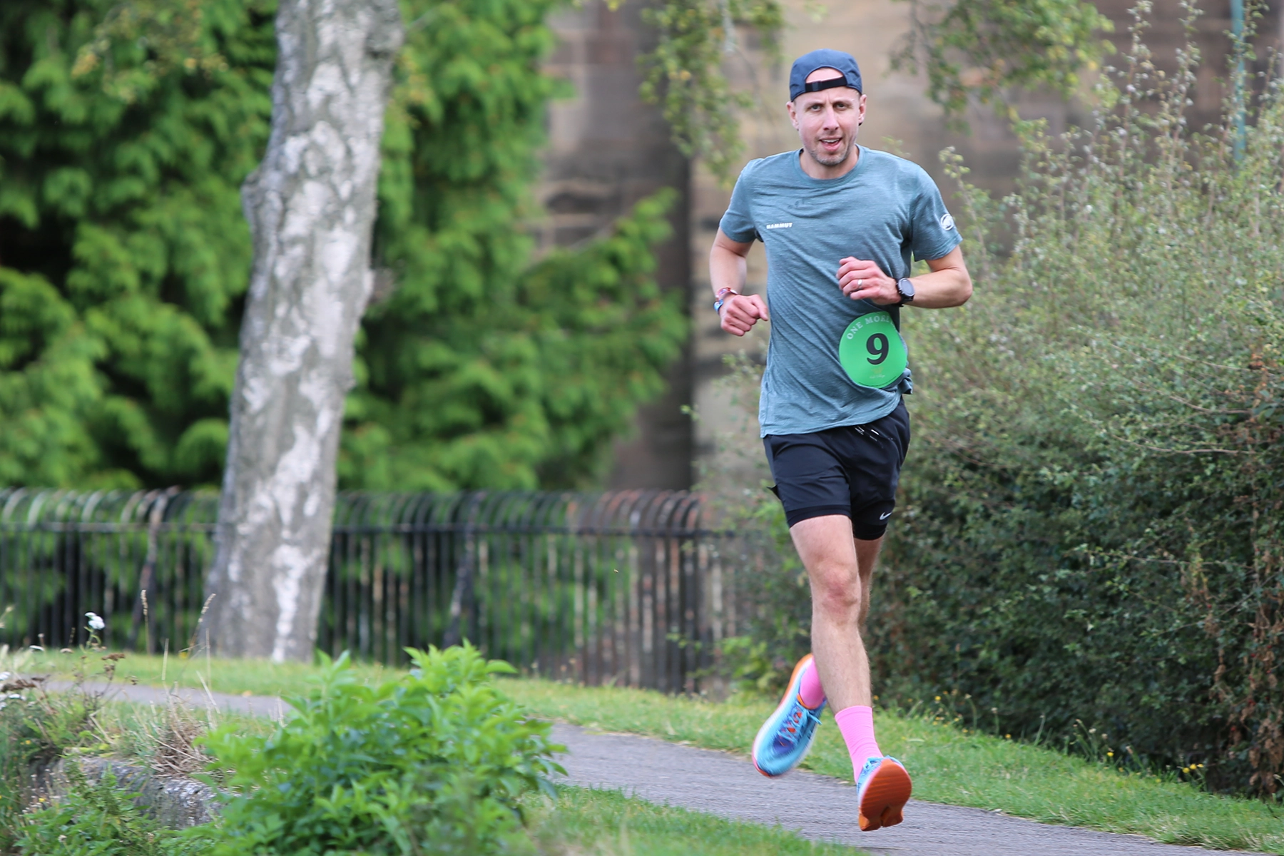 Male runner wearing a backwards cap, green t-shirt, black shorts, and bright pink socks, jogging on a path with a green race bib numbered 9, surrounded by trees and bushes in a park setting.