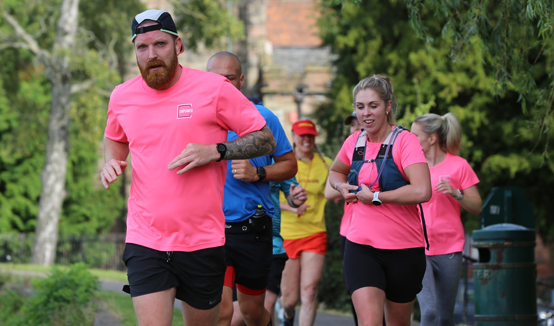 Group of runners in bright pink shirts jogging along a park path, led by a bearded man with a backward cap and a woman adjusting her running vest, with more participants following behind.